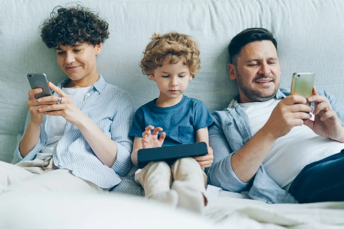 Pai, mãe e filho no sofá, cada um olhando a própria tela.