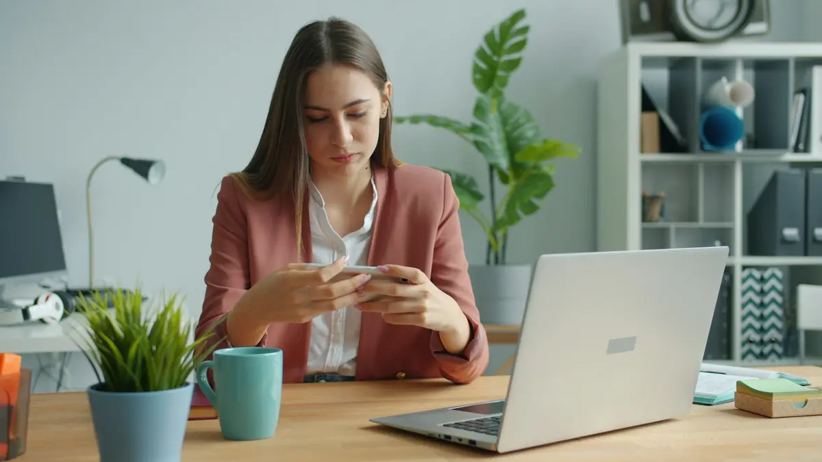 Mulher à mesa de trabalho olha o celular enquanto o notebook espera.